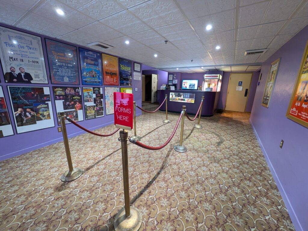 Interior of a theater lobby with a purple color scheme, red rope stanchions, and posters on the wall. A sign reads “Line Forms Here” leading to a concession stand.