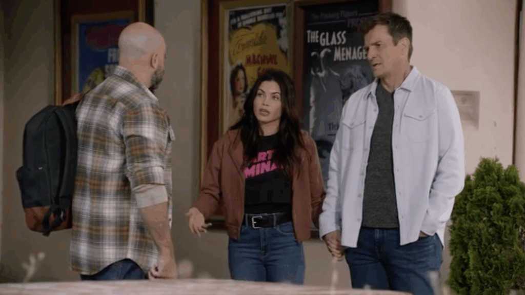 Three people stand talking outside a theater entrance, with movie posters visible behind them. One woman gestures while speaking; the two men listen, one holding her hand.